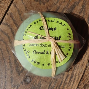 May include: A green round bar of soap wrapped in clear plastic and tied with a brown string. The label reads "Savon artisanal S.A.F. Creme d'avocat Savon doux et  Avocat & 100%  Hyd ate 10: it.  vegeta"