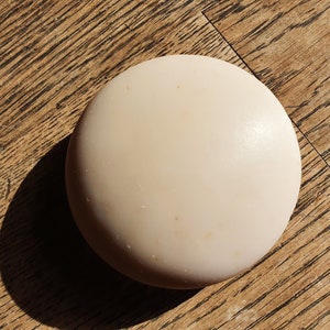 May include: A round, off-white, solid bar of soap on a wooden surface.