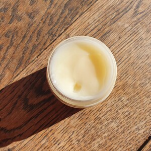 May include: A small, clear jar filled with a pale yellow cream or lotion. The jar is sitting on a wooden surface.