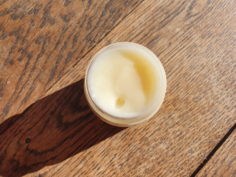 May include: A small, clear jar filled with a pale yellow cream or lotion. The jar is sitting on a wooden surface.