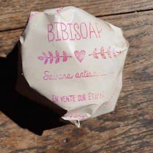 May include: A wrapped bar of soap on a wooden surface. The packaging is light brown with pink text that reads "BIBISOAP" above a heart and leaf design. The text "Savons artisanaux" is also visible.