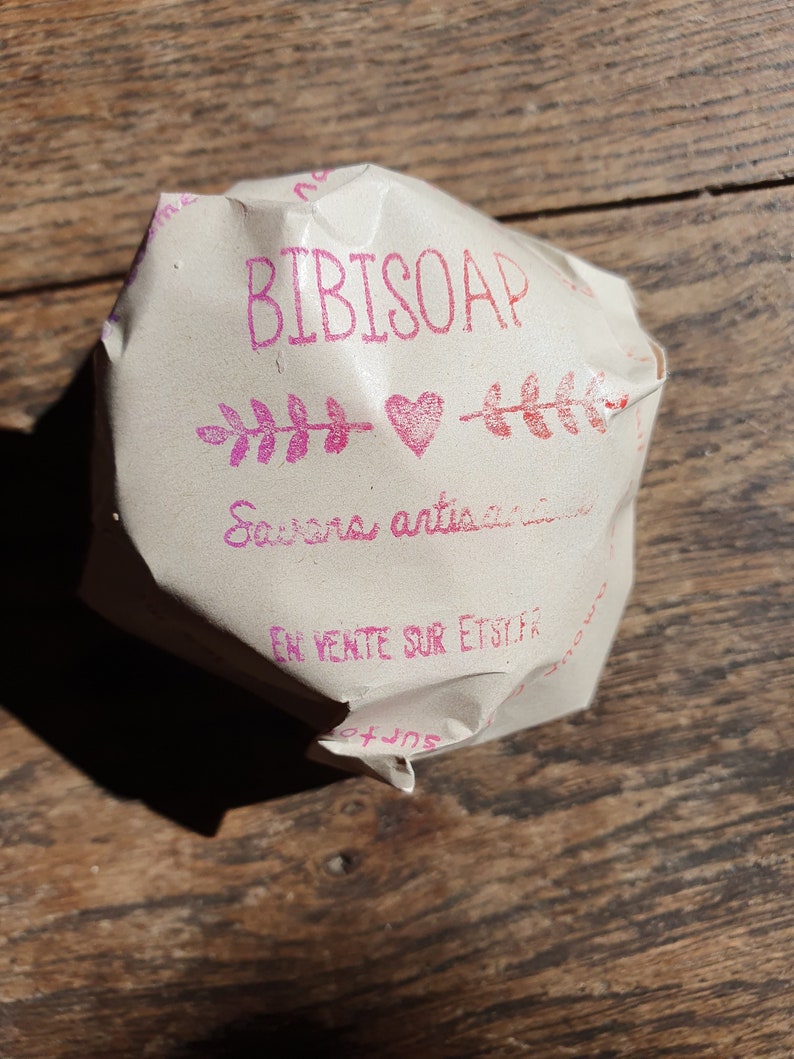 May include: A wrapped bar of soap on a wooden surface. The packaging is light brown with pink text that reads "BIBISOAP" above a heart and leaf design. The text "Savons artisanaux" is also visible.