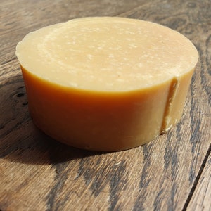 May include: A round, light brown bar of soap. The soap has a smooth, slightly textured surface.