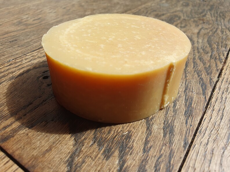 May include: A round, light brown bar of soap. The soap has a smooth, slightly textured surface.