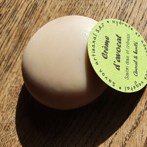 May include: A round, light brown bar of soap with a green label that reads "Cr&egrave;me d'avocat Savon artisanal S.A.F. Savon doux et cr&eacute;meux Avocat & karit&eacute; Hydrate 100% v&eacute;g&eacute;tal"