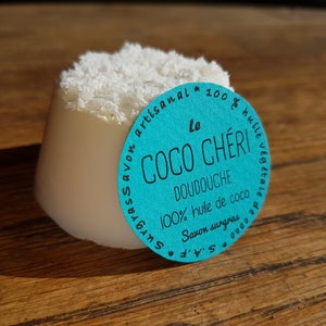 May include: A white, cylindrical soap with a shredded coconut topping. A turquoise label with white text reads "Le Coco Ch&eacute;ri Doudouche 100% huile de coco Savon surgras". The soap is on a wooden surface.