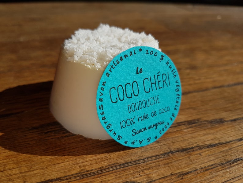 May include: A white, cylindrical soap with a shredded coconut topping. A turquoise label with white text reads "Le Coco Ch&eacute;ri Doudouche 100% huile de coco Savon surgras". The soap is on a wooden surface.