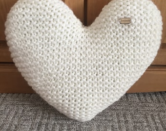 Peut inclure: Un coussin en forme de cœur, tricoté en blanc cassé. Le coussin présente une texture en tricot épais et une petite étiquette en bois avec l'inscription "handmade". Le coussin est posé sur un tapis gris devant un meuble en bois.