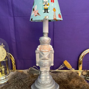 May include: A silver-colored lamp in the shape of a nutcracker soldier. The lamp has a light blue lampshade with a repeating pattern of nutcrackers, ballerinas, and gifts. The lamp is plugged in and ready to use.