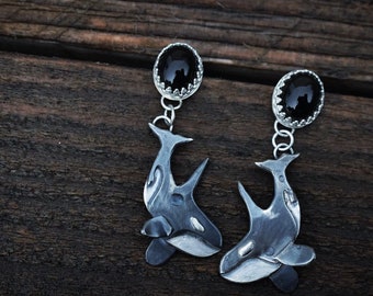 May include: A pair of silver earrings featuring a black onyx stone and a detailed orca whale design.
