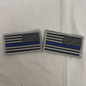 May include: Two rectangular patches featuring a stylized American flag design. The flag has gray and black stripes, a field of black stars, and a blue line. The patches are likely for clothing or accessories.