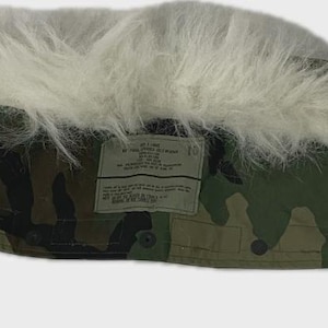 May include: A camouflage-patterned garment with a fluffy, white fur trim. The garment features a green, brown, and black camouflage design, with the white fur adding a contrasting texture and color.