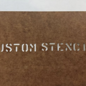 May include: A brown stencil with the words "CUSTOM STENCIL" cut out in a sans-serif font. The stencil is made of a thick, durable material, and the text is centered. The background is white.