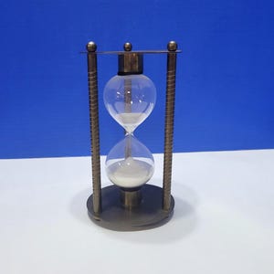 May include: An hourglass with a clear glass bulb filled with white sand, set in a bronze-colored metal frame. The frame has vertical bars with a textured design. The hourglass is a classic timekeeping device.