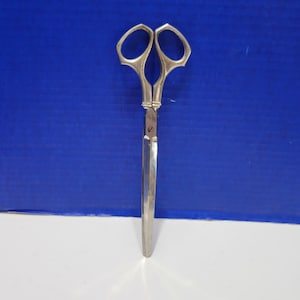 May include: Silver-toned scissors with long, pointed blades and decorative handles. The scissors are closed and are on a white surface.