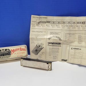 May include: A Hohner Marine Band harmonica, metal case, and instructions. The harmonica is silver and black. The case is off-white with a red and black graphic and the words "Marine Band" and "M. Hohner".