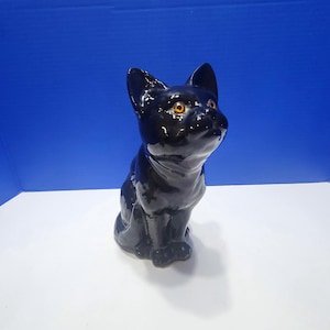 May include: A glossy black ceramic cat figurine with orange eyes. The cat is in a seated position, looking upwards. The figurine has a smooth, reflective surface and is set against a blue and white background.