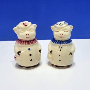 Puede incluir: Un par de figuras de cerdos de cerámica de color crema colocadas una al lado de la otra. El cerdo de la izquierda lleva un sombrero y un cuello de color granate con botones oscuros. El de la derecha lleva un sombrero y cuello azul estampado.