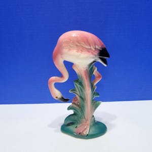 Puede incluir: Figura de flamenco de cerámica, rosa con pico negro y detalles blancos. Se apoya sobre una base verde con hojas estilizadas. La figura tiene un acabado brillante, sobre un fondo azul.