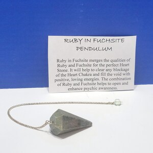 May include: A faceted, gray-green Ruby in Fuchsite pendulum with a silver chain and a clear crystal bead. A white card with the text "RUBY IN FUCHSITE PENDULUM" and description is in the background.