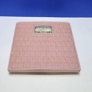 May include: A square bathroom scale in a dusty rose pink color featuring a textured, woven-look surface pattern. The scale includes a central rectangular analog display window at the top, showing numerical weight markings on a rotating internal dial.