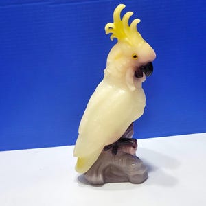 May include: A carved figurine of a cockatoo bird, predominantly off-white with yellow crest feathers and a black beak. The bird is perched on a gray, textured base. The background is a gradient of blue and white.