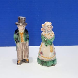 Puede incluir: Dos figuras de cerámica de un hombre y una mujer. El hombre lleva un sombrero de copa, una chaqueta verde y pantalones a rayas. La mujer lleva un gorro, un chal verde y un delantal floral. Las figuras están pintadas con colores brillantes.