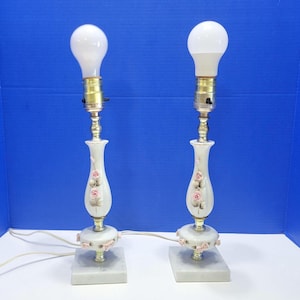 May include: A pair of tall table lamps featuring white ceramic bodies with hand-painted pink rose floral accents. Each lamp has a square marble base, brass-toned metal hardware, and a white light bulb attached to the top socket fixture.