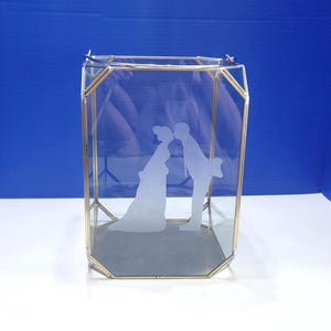 May include: A clear glass terrarium with a gold-colored metal frame. The glass features an etched design of a bride and groom kissing. The terrarium has a geometric shape and is suitable for weddings or home decor.