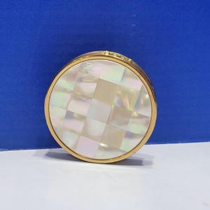 May include: A round compact mirror with a gold-toned metal frame and a lid featuring a mosaic pattern of iridescent, square mother-of-pearl tiles. The surface displays a subtle pearlescent sheen with varying shades of white, cream, and soft pink.