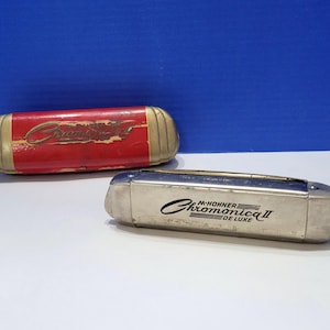 Pode incluir: Duas harmónicas, uma num estojo vermelho com detalhes dourados e a palavra "Chromonica II". A outra harmónica é prateada com o texto "M. Hohner Chromonica II de Luxe". Ambas estão numa superfície branca.