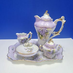 Puede incluir: Un juego de té de porcelana que incluye una tetera, una lechera, un azucarero con tapa y un joyero con tapa sobre una bandeja rectangular. Las piezas son de color morado claro con flores violetas pintadas y bordes dorados.