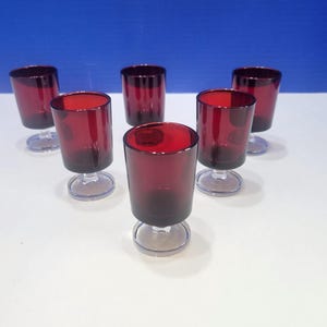 May include: A set of six small drinking glasses with deep red bowls and clear glass stems and circular bases. The glasses are arranged in a triangular formation, featuring a cylindrical shape with a smooth finish and a sturdy footed design.