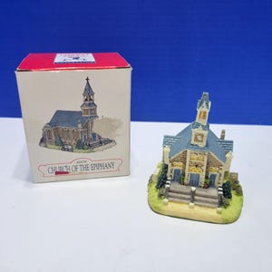 May include: A miniature Church of the Epiphany figurine with a blue roof, stone-textured walls, and a steeple, accompanied by its original cardboard box featuring a matching illustration and the text Church of the Epiphany printed on the front.