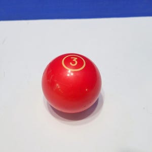 May include: A red billiard ball with the number 3 in a gold circle. The ball is smooth and glossy, and it is set against a white surface with a blue background.