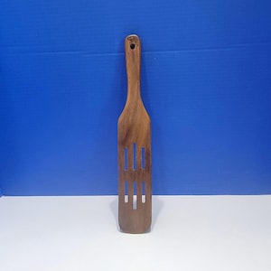 Puede incluir: Una espátula ranurada de madera marrón con un mango largo que presenta un orificio circular en la parte superior para colgar. El utensilio de cocina tiene una cabeza plana y rectangular con tres ranuras verticales cortadas en la madera.