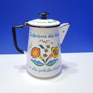 May include: White enamel coffee pot with a black handle and lid. The pot is decorated with colorful floral designs and text in blue. The text reads "Kaffetåren den bästa av alla jordiska drycker."