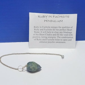 May include: A Ruby in Fuchsite pendulum with a silver chain and a green and blue stone. A white card with the text "RUBY IN FUCHSITE PENDULUM" is in the background. The card describes the stone's properties.