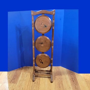 May include: A tall, brown wooden stand featuring three circular, vertically aligned shelves. Each shelf is decorated with a painted fruit motif. The frame consists of two vertical posts connected by horizontal bars, with small floral accents at the top.