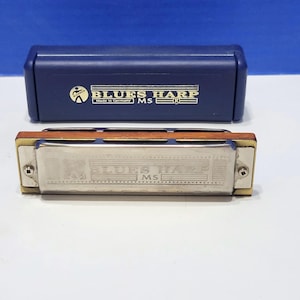 Puede incluir: Una armónica y su estuche azul marino. La armónica es plateada con cuerpo de madera y detalles dorados. El estuche tiene las palabras "BLUES HARP" impresas en dorado.