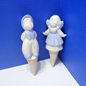 Op de afbeelding: Twee keramische beeldjes van kinderen, een jongen en een meisje, beide met lichtblauwe en witte kleding. De jongen draagt een hoed en het meisje heeft een muts. Beide figuren zijn gemonteerd op beige kegelvormige bases.