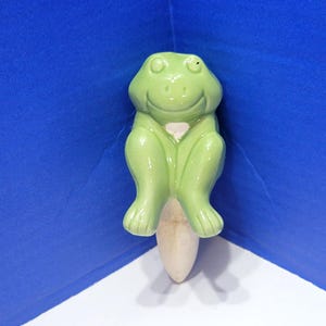 Op de afbeelding: Een lichtgroen keramieken kikkerbeeldje met een lachend gezicht. De kikker zit met gekruiste benen. Het beeldje is bevestigd aan een lichtgekleurde, langwerpige basis. De achtergrond is blauw en wit.