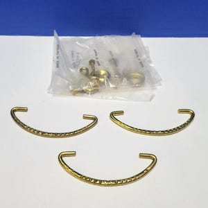 Può includere: Tre maniglie in metallo dorato con un motivo decorativo. Le maniglie sono curve e hanno una finitura liscia.