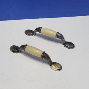 May include: Two cabinet pulls featuring a central cream-colored cylindrical insert flanked by metallic, dark-toned curved ends. Each handle has a rounded base at both attachment points, showcasing a weathered, aged finish on the metal components.