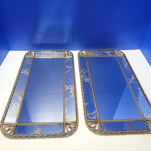 Op de afbeelding: Twee rechthoekige spiegels met blauw glas en goudkleurige metalen frames. De frames hebben een touwachtige rand en decoratieve hoekelementen. De spiegeloppervlakken hebben een goud-wit marmerpatroon.