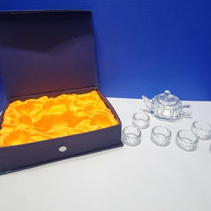 May include: A clear glass tea set with a teapot and six small cups. The teapot and cups have a faceted design. A dark purple box with a yellow satin lining is open next to the tea set.