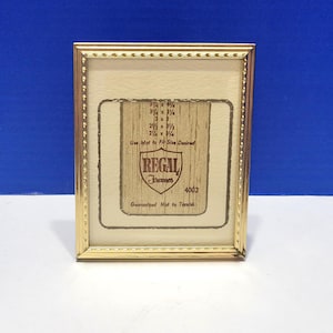 May include: A rectangular gold-colored picture frame with a cream-colored interior. The frame has a decorative beaded edge. Inside the frame is a tan-colored insert with text and measurements. The text includes the brand name "REGAL" and the phrase "Guaranteed Not to Tarnish."
