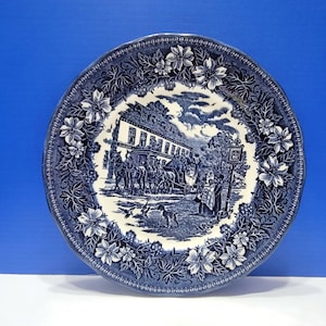 May include: A blue and white decorative plate with a floral border. The center features a detailed scene of a building, people, and horses. The plate has a diameter of approximately 10 inches.