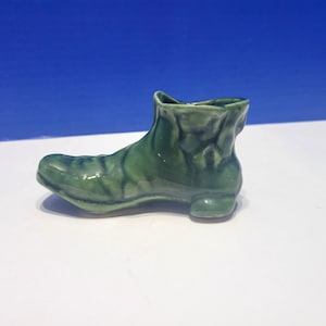 May include: A small, ceramic green boot figurine. The boot has a glossy finish and is detailed with textured patterns. The boot is a miniature decorative item, likely for display or collection. The background is a gradient of white and blue.