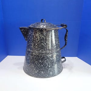 May include: A black enamel coffee percolator featuring a white speckled pattern throughout the exterior. The pot includes a matching lid, a side handle with a dark wooden grip, a wide spout, and a metal bail handle attached to the top rim.